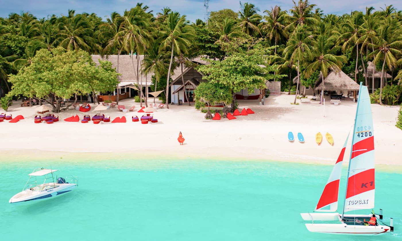 a view from the sea to the beach. The sea is turquise blue, the beach has various watersport items on it and a woman in a red dress at the shoreline.  in the background are palm trees