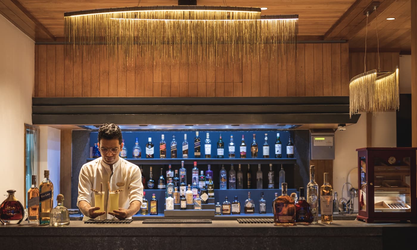 A barman wearing his uniform stood behind a car.  behinfd him are shelves with different bottles on them