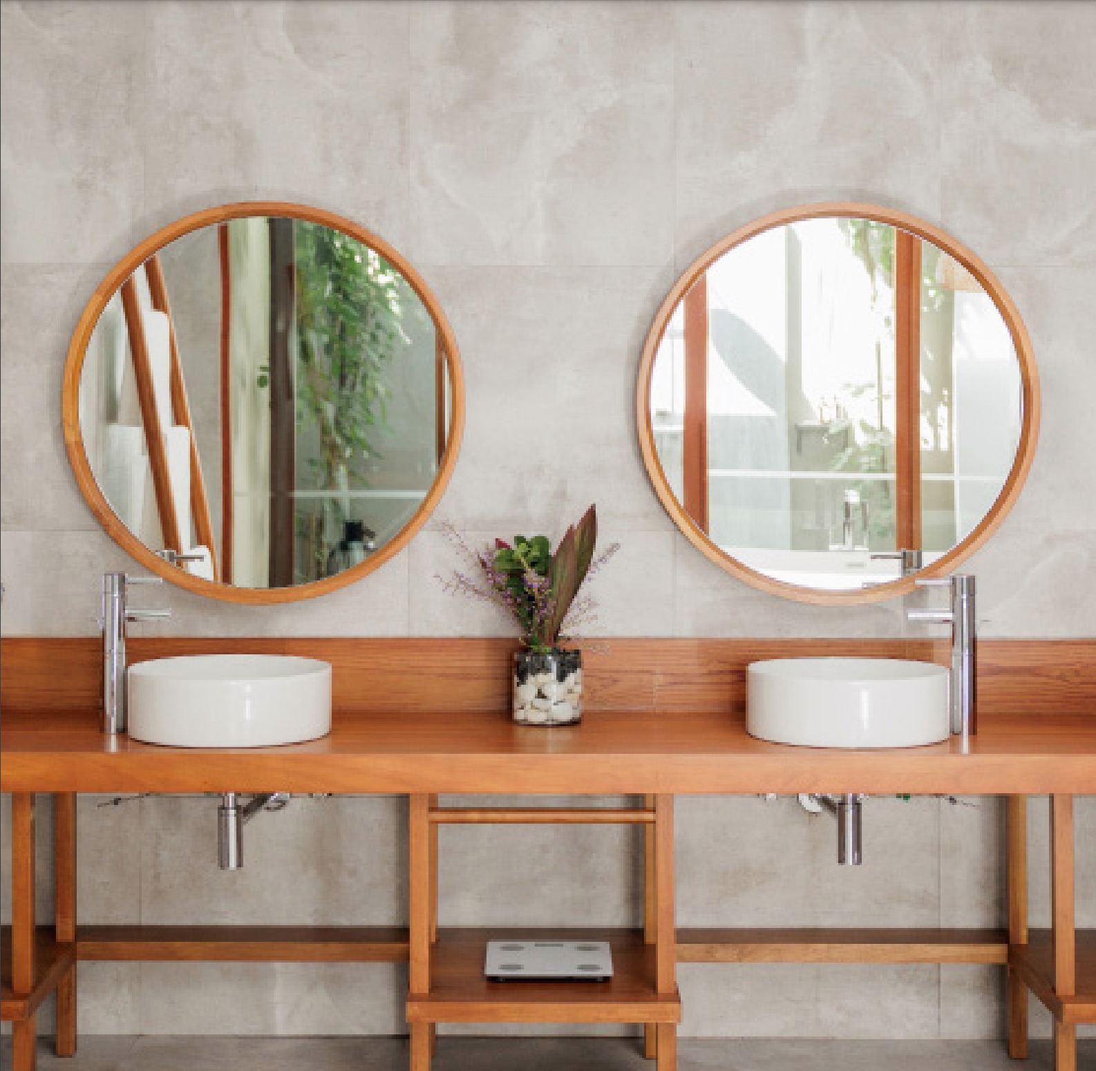 A double washbasin unit in the bathroom of a villa.  There are spaces underneath the basins for a wheelchair to roll under.  The unit is made of wood, there are round white basins and a plant between them.  On the wall are two large round mirrors