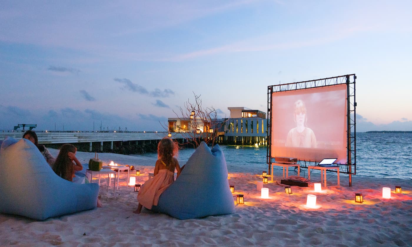 A beach with a woman sat on a bean bag in front of a large cinema screen. She is watching a movie. There are candles dotted around in the sand