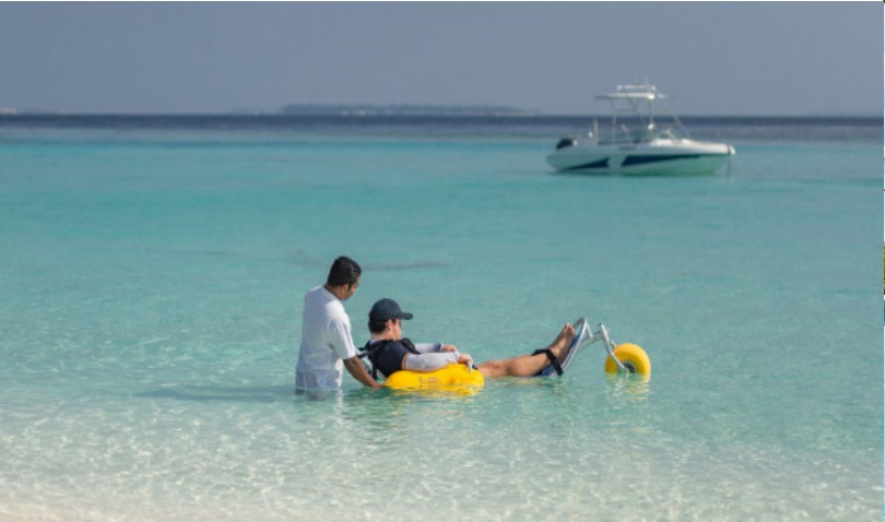 A man in the sea in a floatuing beach wheelchair.  he is being assisted by another man who is holding the back ofthe chair.  In the background is a boat